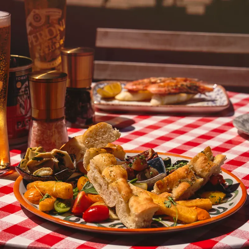 Sun-drenched antipasti spread on red-gingham linen — focaccia, olives, blistered tomatoes, big king prawns and a cold glass of Peroni.
