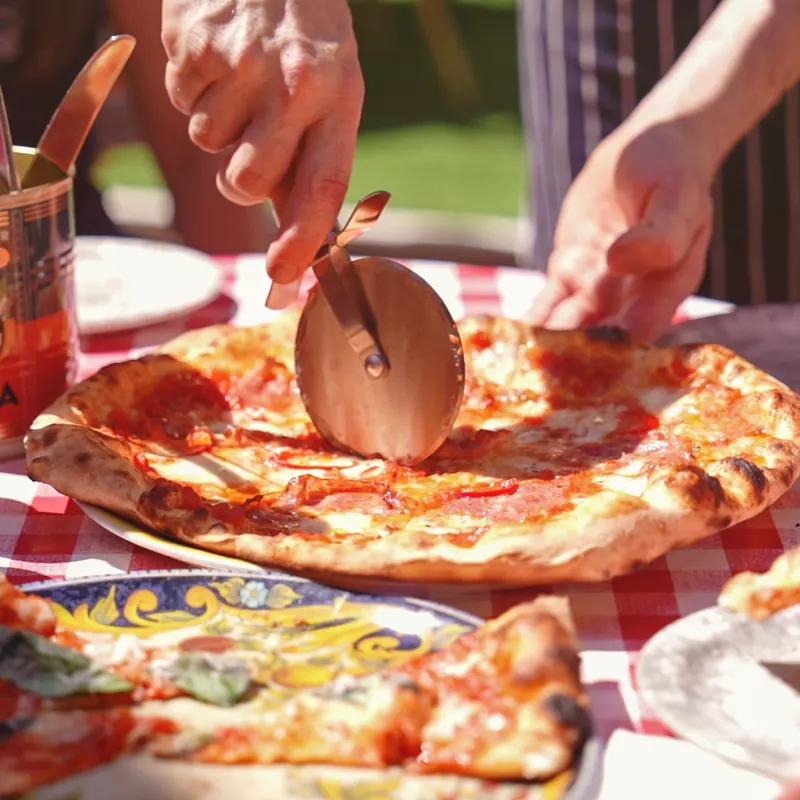A pizzaiolo’s hands slicing a Margherita with a wooden-handled pizza wheel.