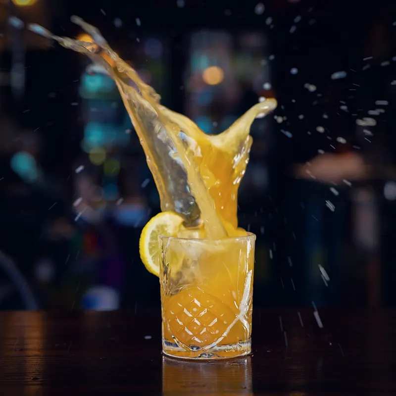 Mid-pour cocktail splashing over a crystal tumbler with a lemon wheel, warm bar lights blurred behind.