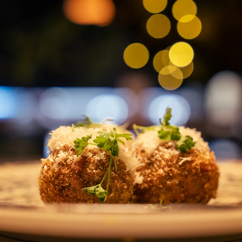 Two golden-crisp arancini balls topped with grated parmesan and microgreens, set against soft bokeh lights.