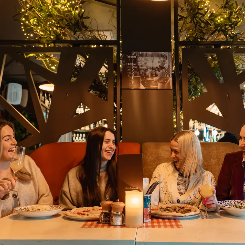 A table of friends laughing over Napoli pizza, antipasti and glasses of wine, with festoon lights warming the room behind them.